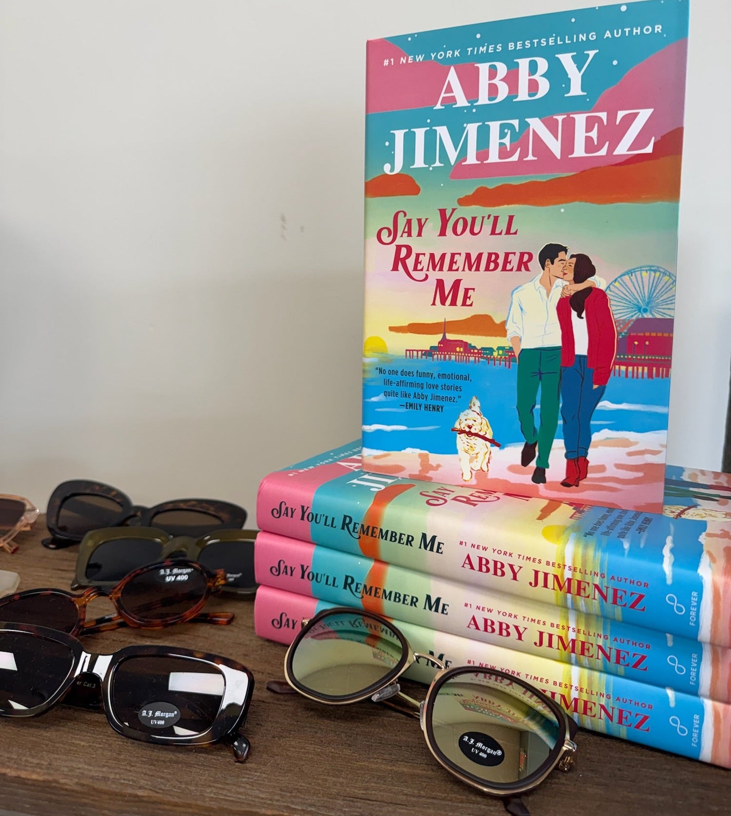 Stack of books titled 'Say You'll Remember Me' by Abby Jimenez with sunglasses on a wooden surface.